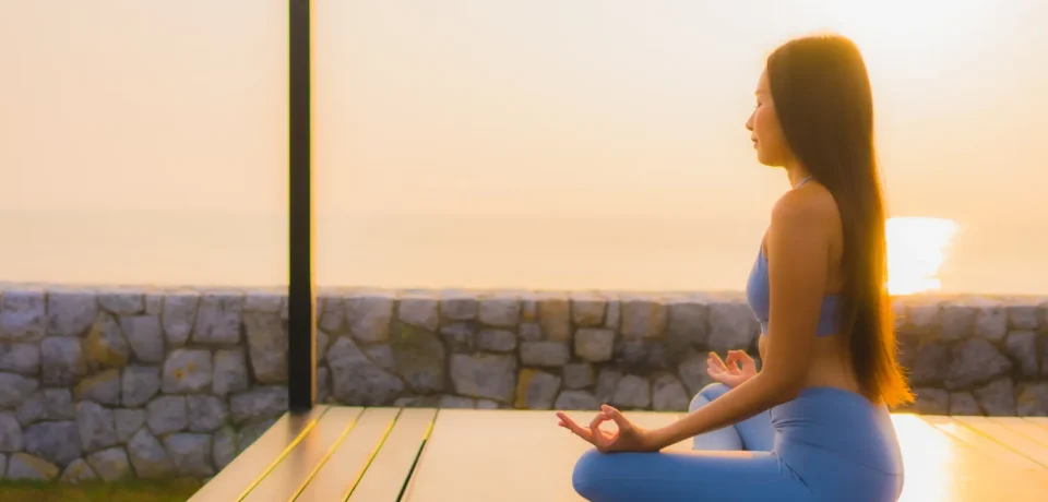 Mujer meditando en una terraza al amanecer, vista al mar, vestida con un conjunto de yoga azul, promoviendo la paz y la tranquilidad.