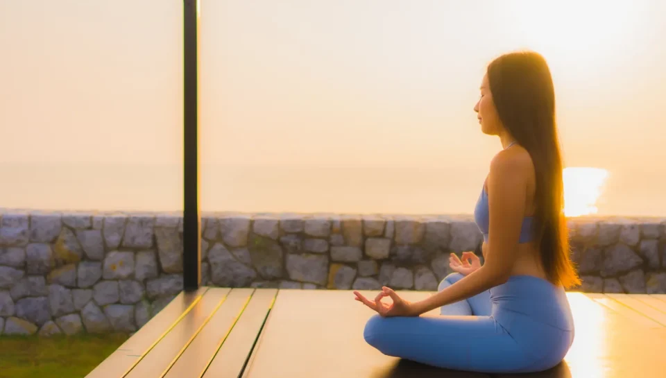 Mujer meditando en una terraza al amanecer, vista al mar, vestida con un conjunto de yoga azul, promoviendo la paz y la tranquilidad.