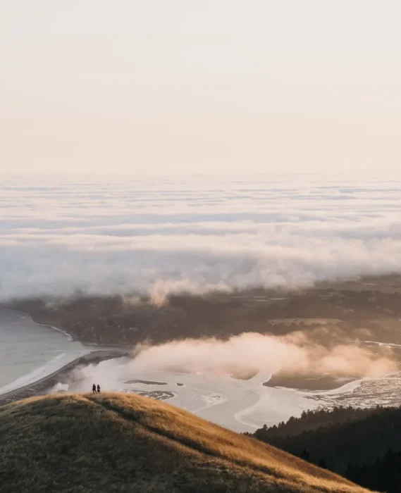 Vista panorámica de un paisaje costero al amanecer, con nubes bajas cubriendo el mar y un río serpenteante en la parte inferior. Dos personas se encuentran de pie en la cima de una colina, rodeadas de vegetación, disfrutando de la naturaleza y la tranquilidad del entorno.