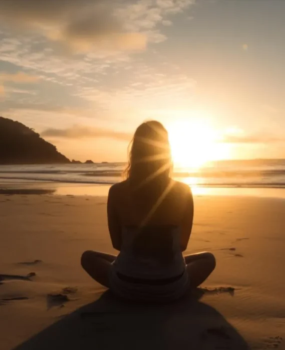 Mujer meditando en la playa al atardecer, con el sol brillando en el horizonte y el mar sereno de fondo.