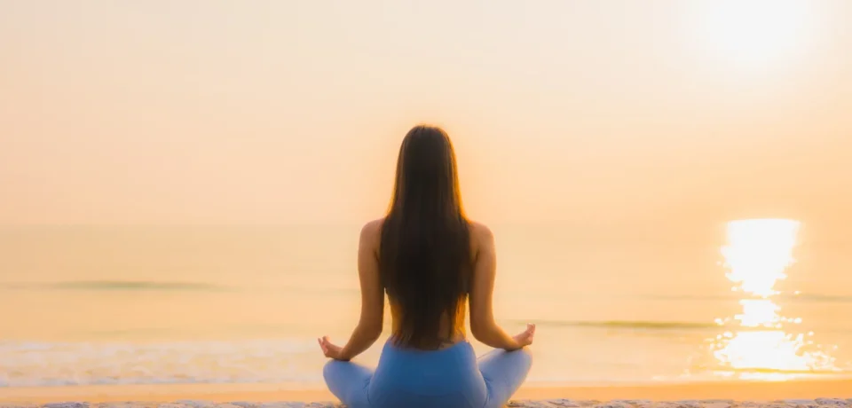 Mujer meditando en la playa al amanecer, sentada sobre una pared de piedra con el mar y el sol reflejándose en el agua.