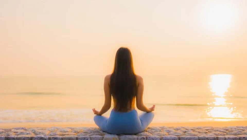 Mujer meditando en la playa al amanecer, sentada sobre una pared de piedra con el mar y el sol reflejándose en el agua.